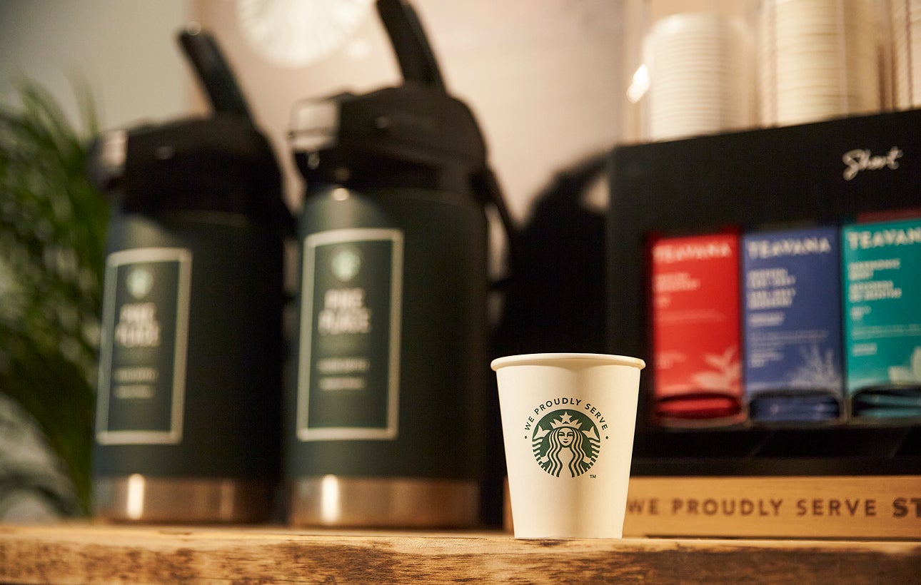 Une tasse en carton portant l’inscription We Proudly Serve Starbucks est posée sur un comptoir en bois, devant de grands dist