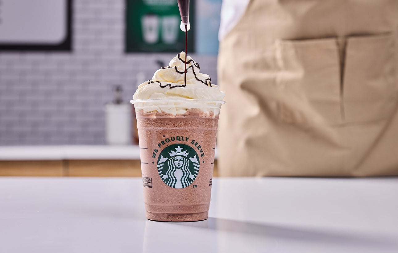 Une boisson We Proudly Serve Starbucks nappée de crème fouettée, avec un filet de chocolat ajouté par un barista.