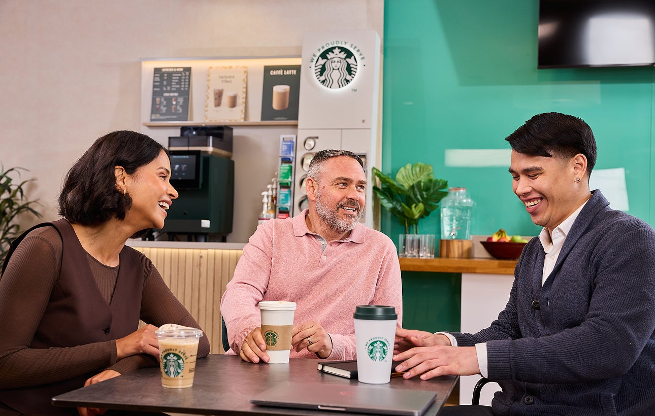 Des collègues assis ensemble avec des tasses We Proudly Serve Starbucks et une machine à café en libre-service en arrière-pla