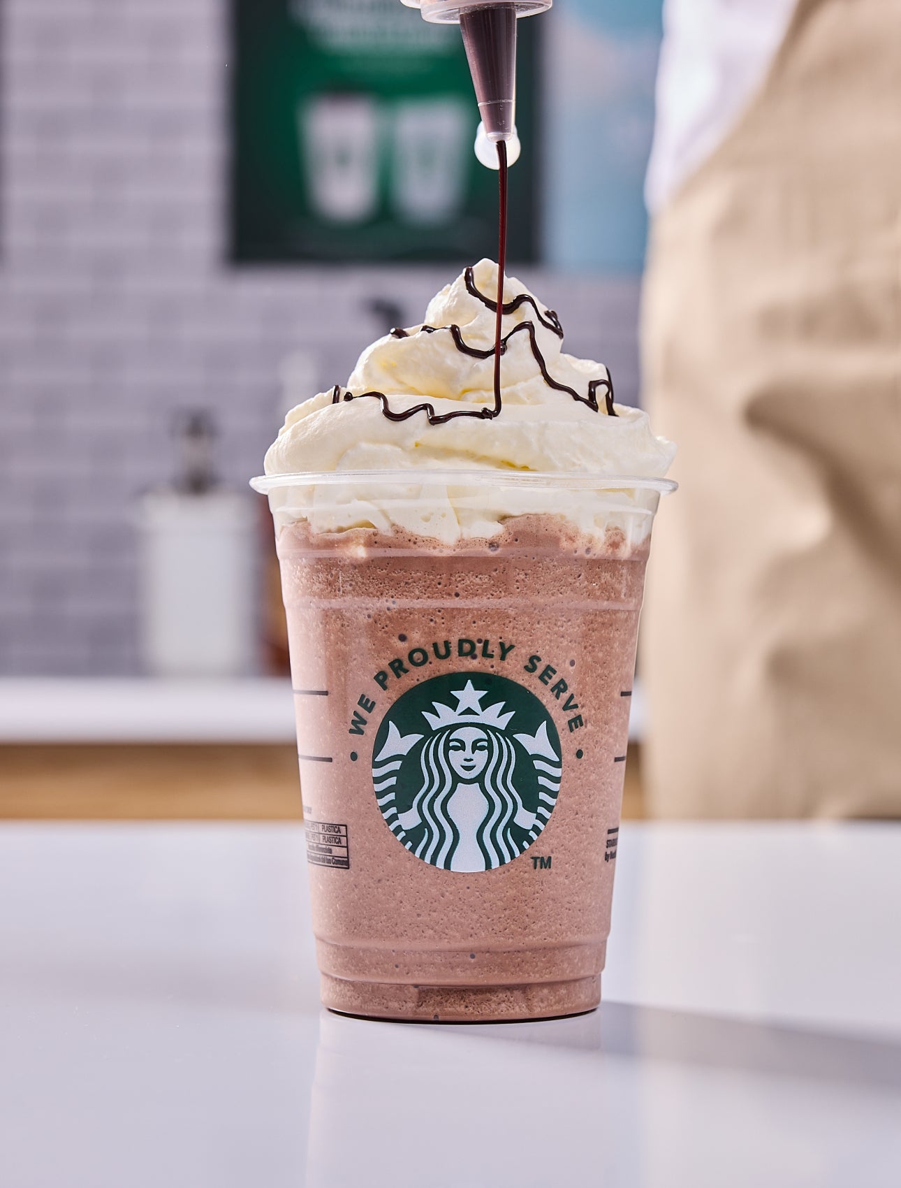 Une boisson We Proudly Serve Starbucks nappée de crème fouettée, avec un filet de chocolat ajouté par un barista.