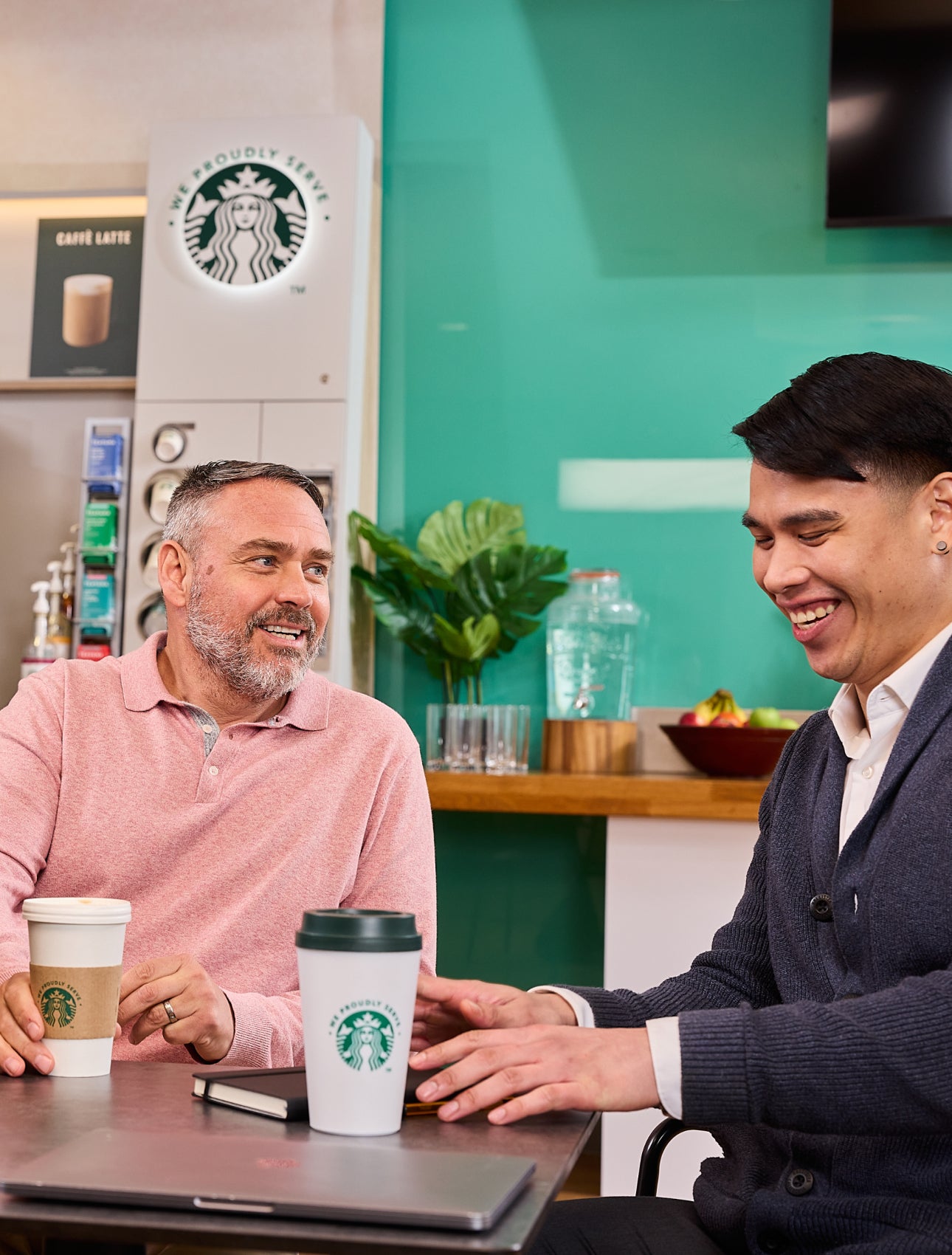 Des collègues assis ensemble avec des tasses We Proudly Serve Starbucks et une machine à café en libre-service en arrière-pla