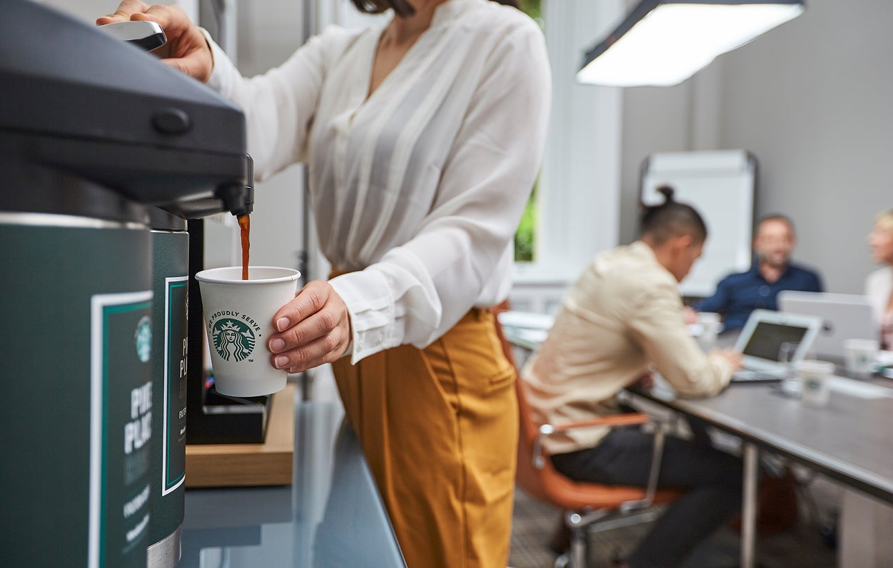 Une personne remplit une tasse en carton We Proudly Serve Starbucks avec du café délivré par un distributeur de café commerci