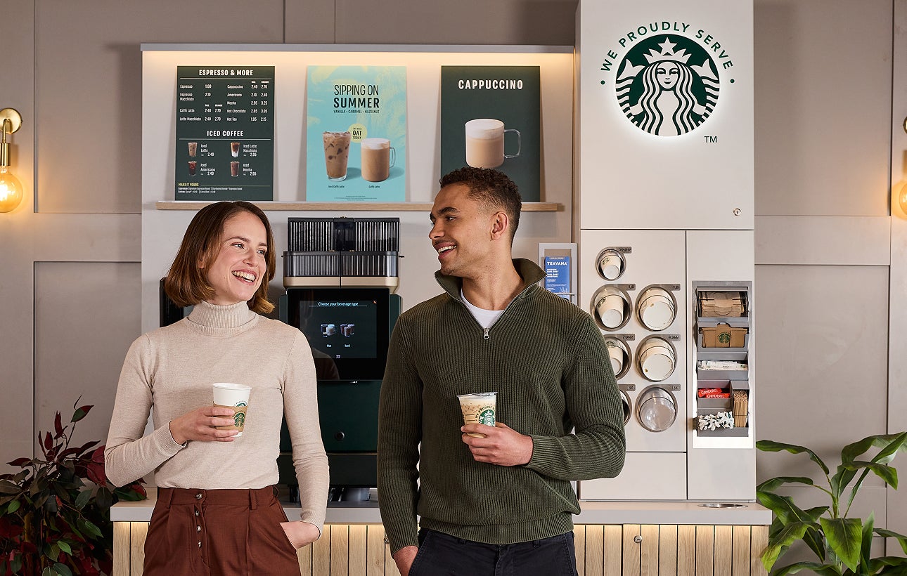 Deux personnes debout, en train de discuter, tenant une tasse de café We Proudly Serve Starbucks devant une station de café e