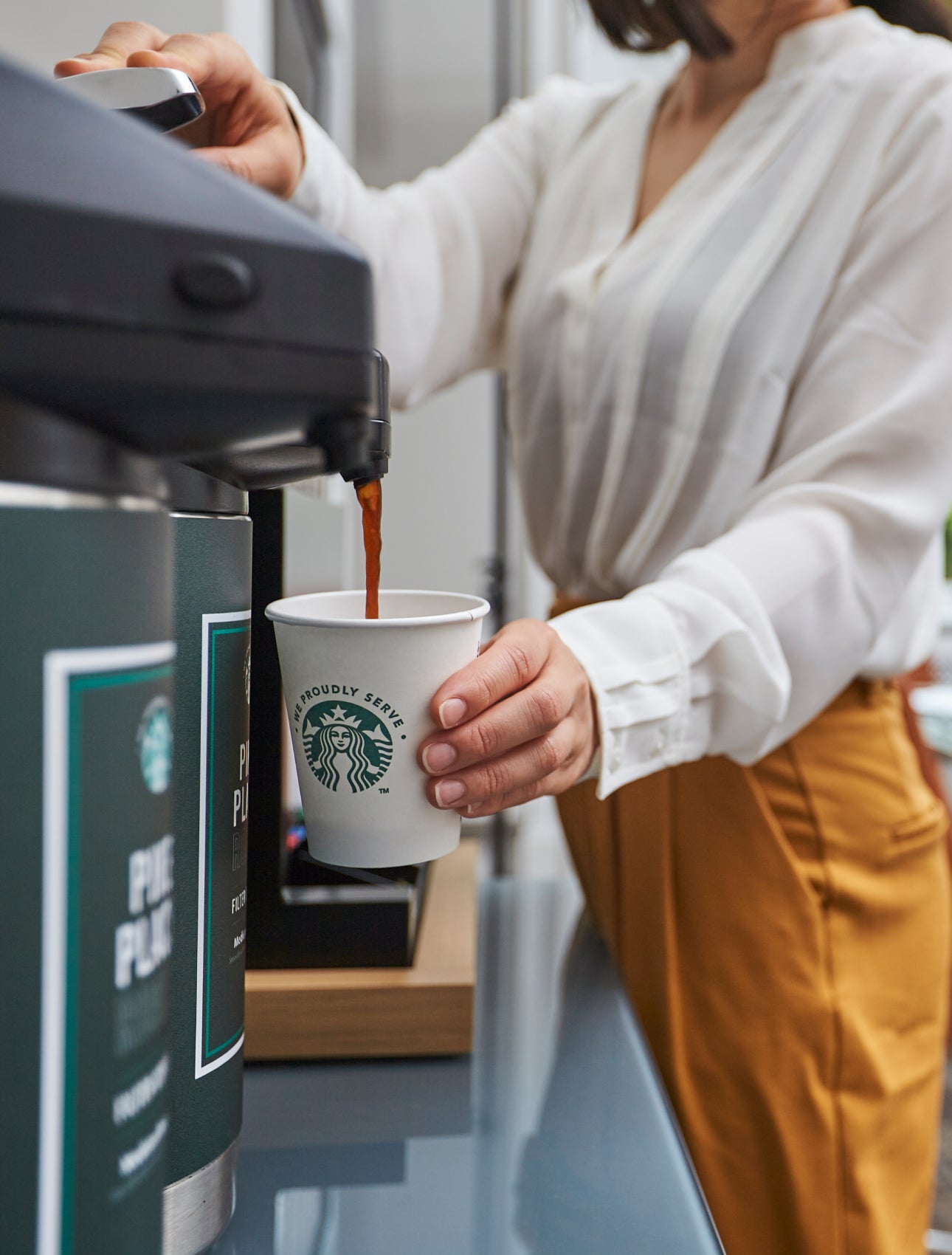 Une personne remplit une tasse en carton We Proudly Serve Starbucks avec du café délivré par un distributeur de café commerci