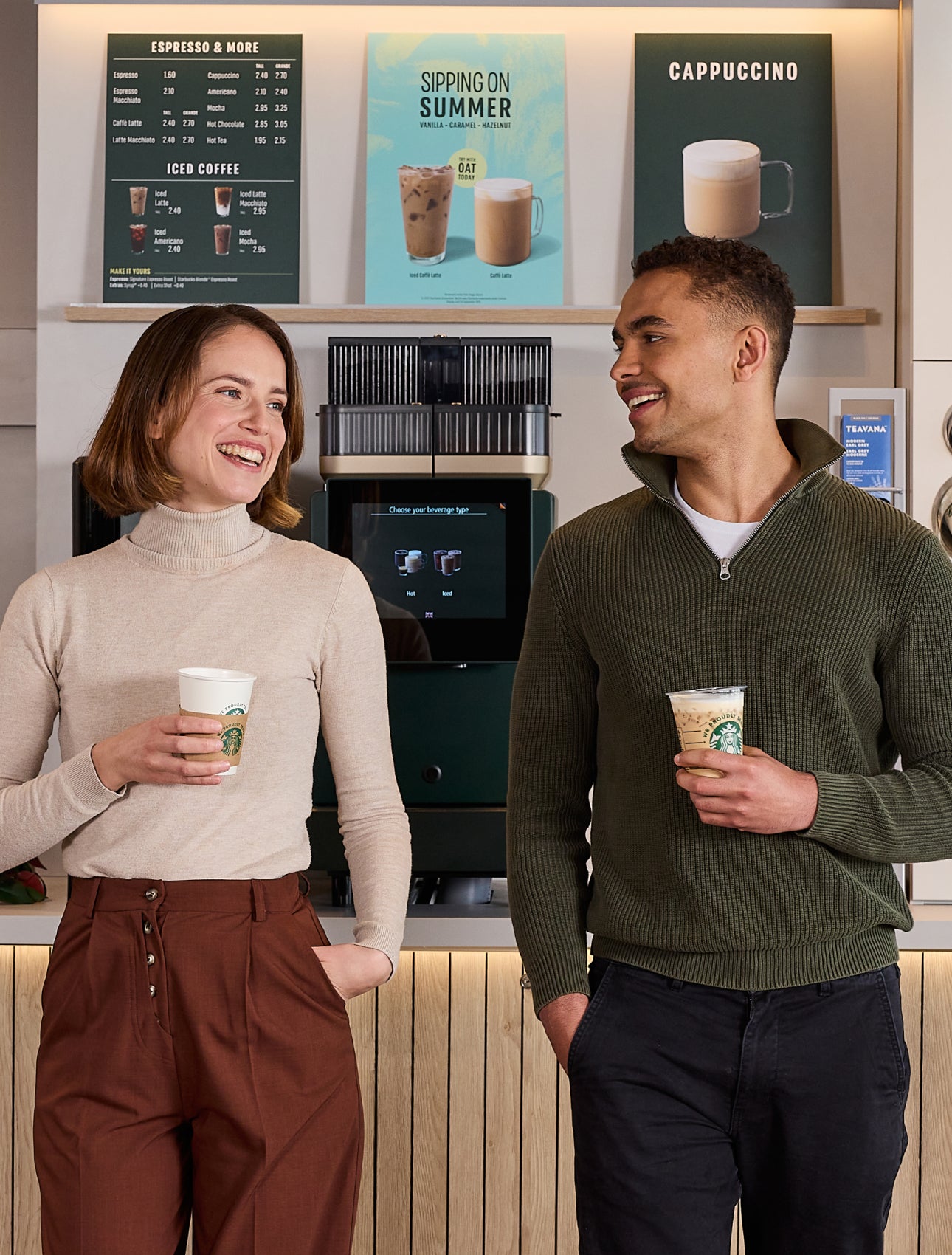 Deux personnes debout, en train de discuter, tenant une tasse de café We Proudly Serve Starbucks devant une station de café e