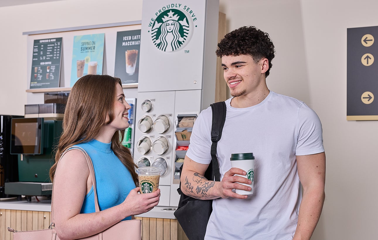 Un homme et une femme debout devant une station à café en libre-service We Proudly Serve Starbucks