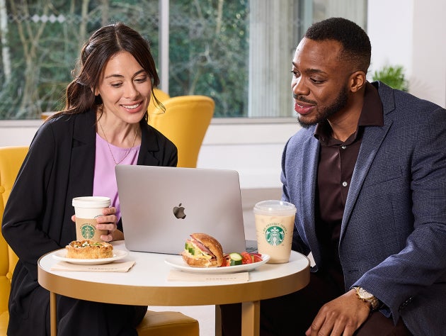 Deux personnes assises devant un ordinateur portable, avec du café We Proudly Serve Starbucks et des sandwichs sur la table