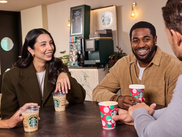 Quatre personnes assises à la table d’un café, souriantes et tenant des boissons Starbucks dans des tasses festives et standa