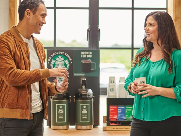 Deux personnes debout, en train de discuter, tenant une tasse We Proudly Serve Starbucks devant des distributeurs de café