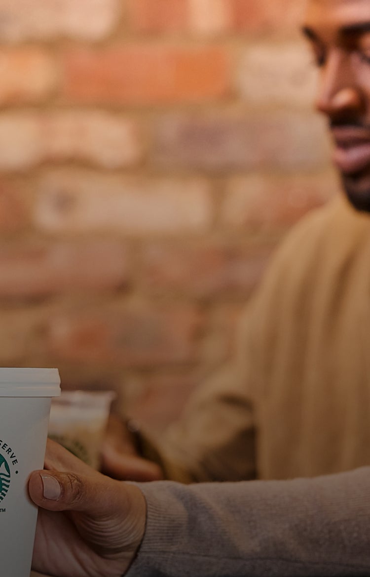 Une personne tenant une tasse We Proudly Serve Starbucks devant un fond de briques flou.
