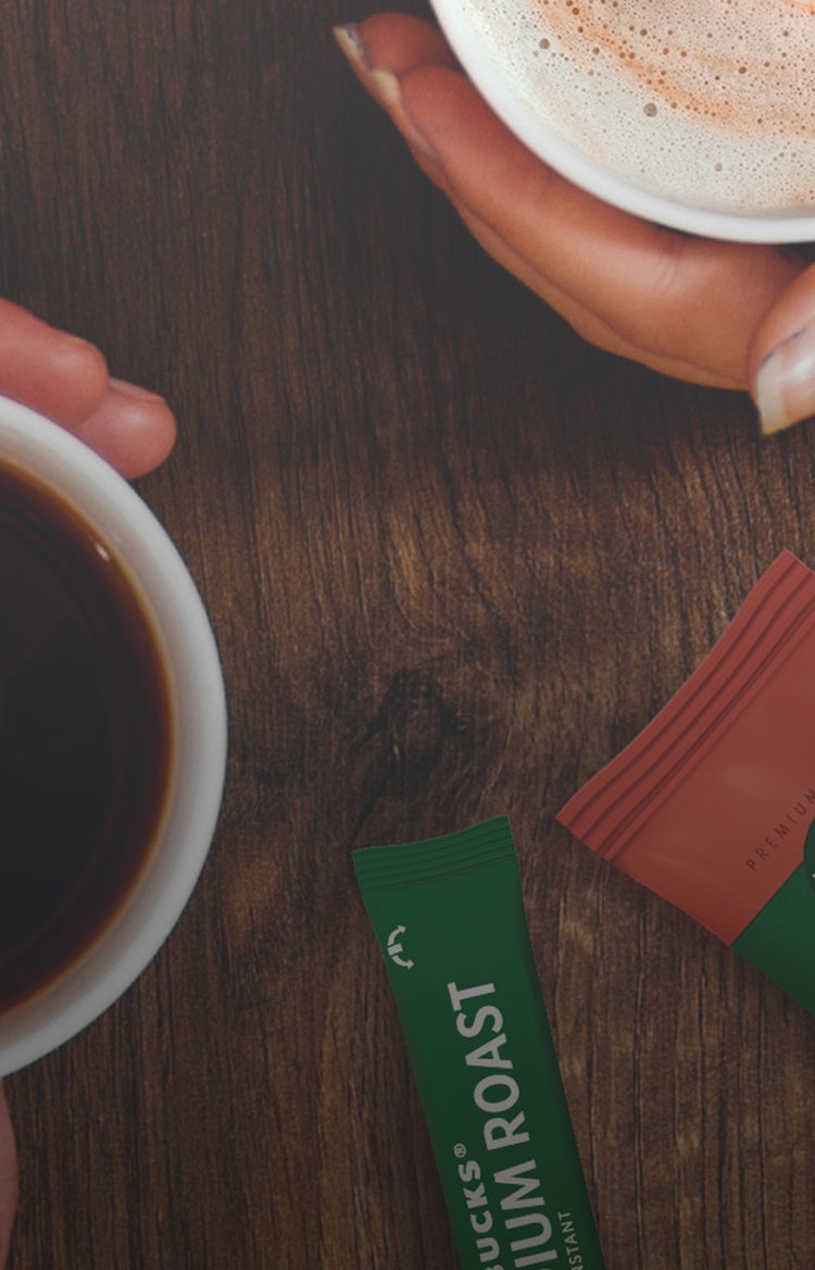 Deux mains tiennent un café noir et un cappuccino mousseux sur une table, à côté de sachets de café instantané Starbucks® Pre
