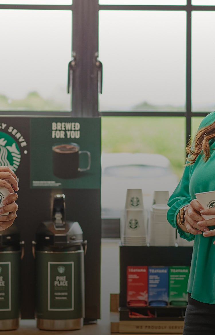 Deux personnes debout, en train de discuter, tenant une tasse We Proudly Serve Starbucks devant des distributeurs de café