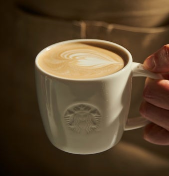 Une main tenant une tasse blanche Starbucks remplie d’un latte, nappé d’un dessin en forme de cœur