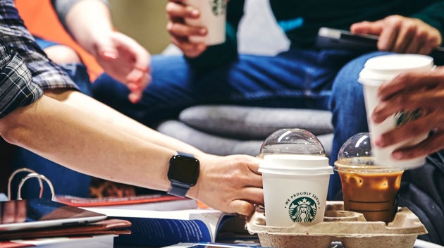 Un groupe d’étudiants assis autour d’une table sur laquelle se trouvent des livres et des cahiers, l’un d’eux prend un café W